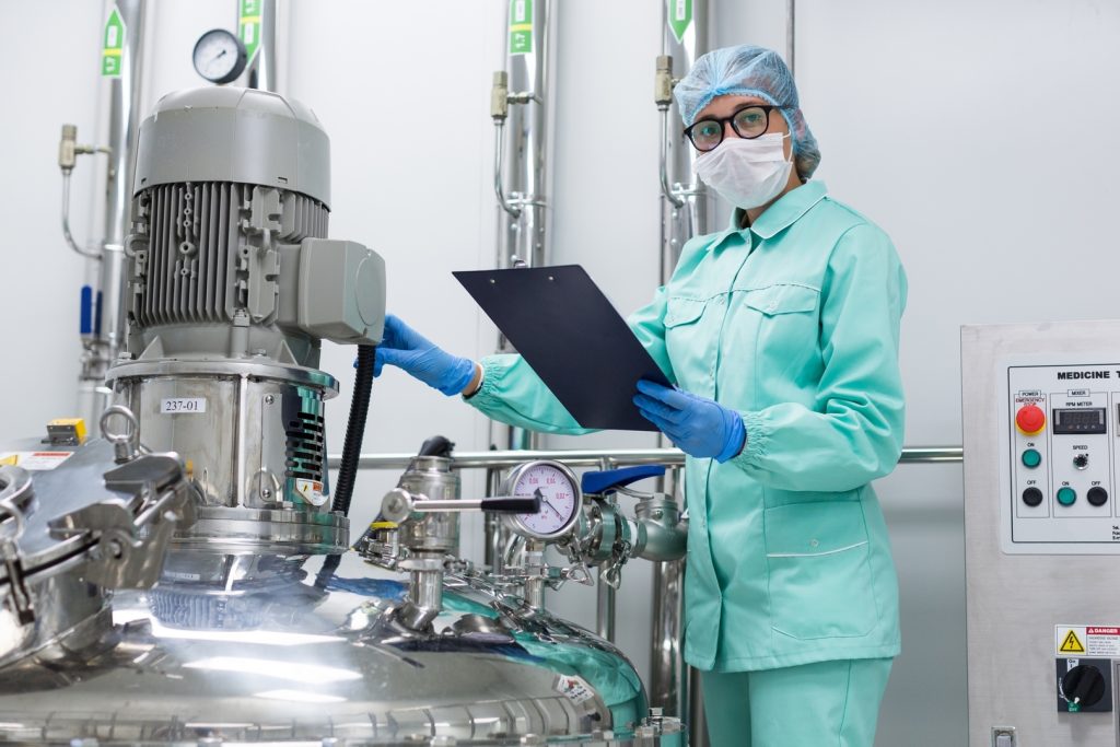scientist in blue lab uniform stand near big pressure tank , hold tablet , look at camera